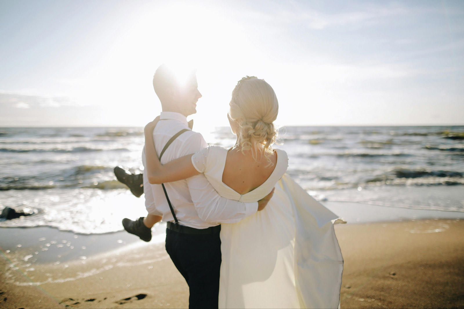 Couple on a sunny beach. The man is carrying the woman across the beach. They are both dressed in wedding attire.